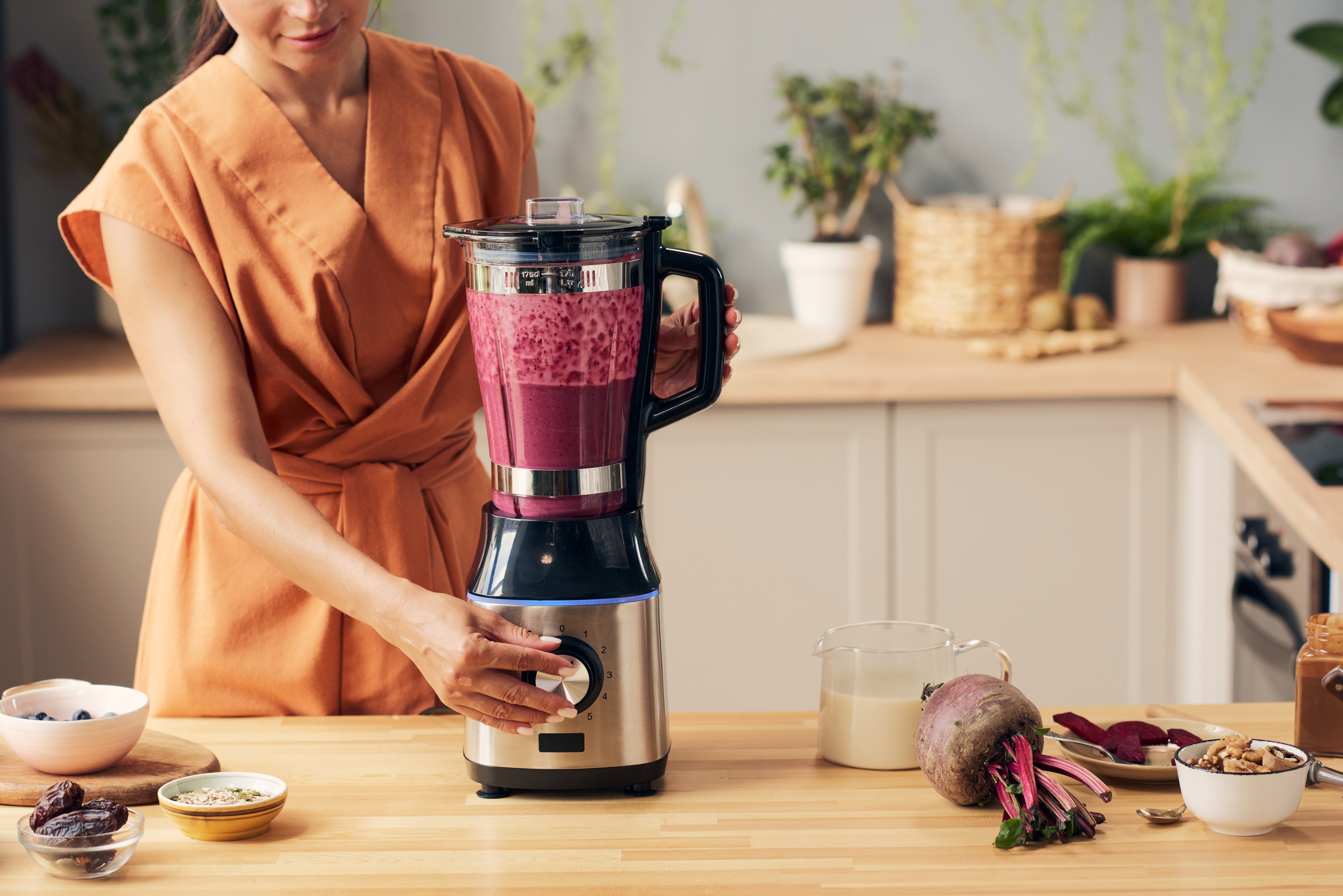 Young housewife with electric blender preparing beetroot smoothie by kitchen table with bowls containing ingredients Young housewife with electric blender preparing beetroot smoothie by kitchen table with bowls containing ingredients