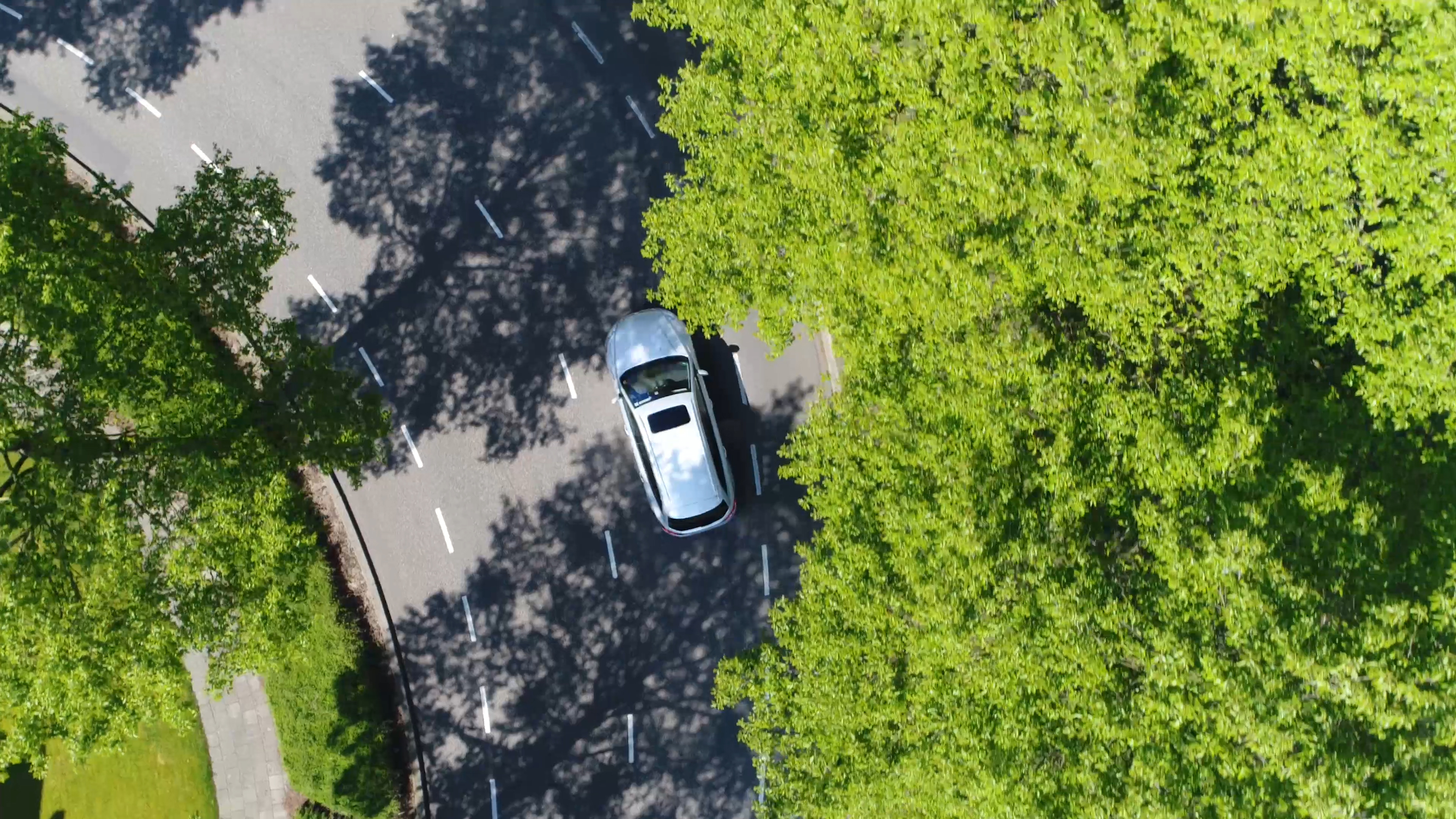 Aerial following car top-down view this grey colored station wagon is driving over two way street corner green trees on both sides of street Aerial following car top-down view this grey colored station wagon is driving over two way street corner green trees on both sides of street