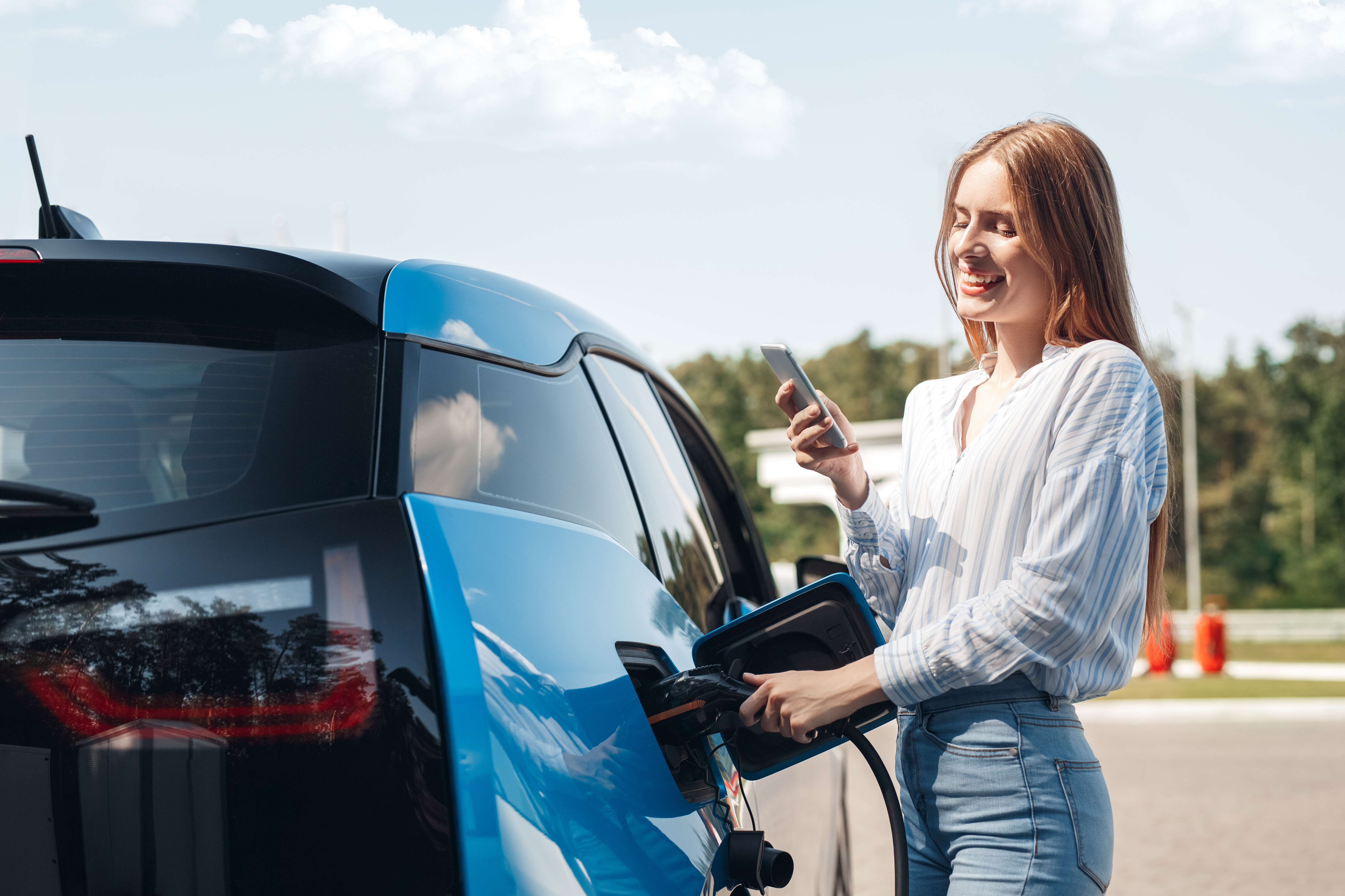 Young beautiful woman traveling by electric car having stop at charging station standing plugging cable browsing internet on smartphone smiling joyful while charing Young beautiful woman traveling by electric car having stop at charging station standing plugging cable browsing internet on smartphone smiling joyful while charing